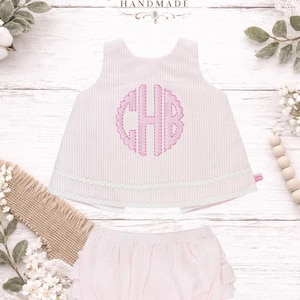 Pode incluir: Conjunto de roupa de bebé às riscas rosa e branco. A parte de cima tem um monograma bordado a rosa e uma borda com vieiras. Os bloomers a condizer têm bordas com folhos. O texto "Kenton Co Handmade" está presente.