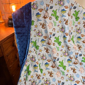 May include: A blue and white blanket featuring a pattern of cartoon characters. The blanket has a soft, plush blue backing. The pattern includes images of a cowboy, a space ranger, and other characters on a light blue background.