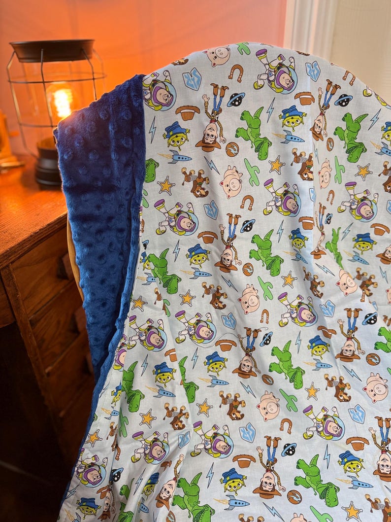 May include: A blue and white blanket featuring a pattern of cartoon characters. The blanket has a soft, plush blue backing. The pattern includes images of a cowboy, a space ranger, and other characters on a light blue background.