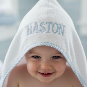 May include: A white hooded baby towel with the name "HASTON" embroidered in light blue. The towel has a light blue trim and is wrapped around a smiling baby.