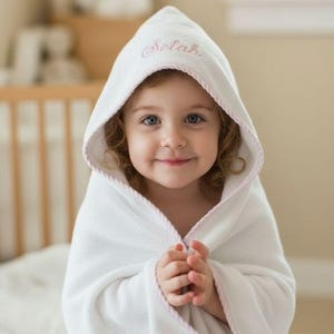 May include: A white hooded baby towel with pink trim and the name "Selah" embroidered on the hood. The towel is wrapped around a child. The background includes a cot and a window.