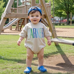 Puede incluir: Un niño pequeño con un body beige con el nombre "PARKER" en letras azules y verdes. El niño lleva una gorra de béisbol azul y zuecos azules. Un columpio de madera se encuentra en el fondo.