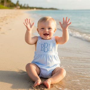 Puede incluir: Un bebé con un pelele azul claro con las letras bordadas "RBL" está sentado en una playa de arena, levantando ambas manos. El niño sonríe, con el océano y las palmeras al fondo.