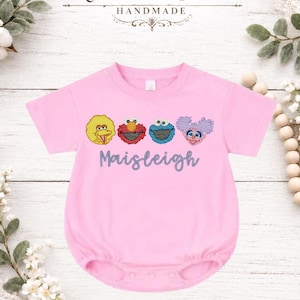 Op de afbeelding: Roze babyromper met geborduurde Sesamstraat-personages: Big Bird, Elmo, Koekiemonster en Abby Cadabby. De naam "Maisleigh" is onder de personages geborduurd. Het merk "Kenton & Cotton Handmade" staat bovenaan.