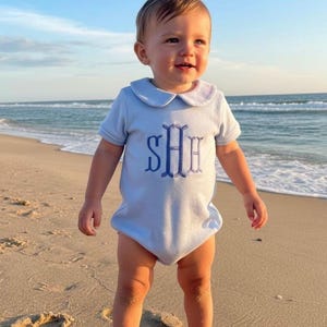 Op de afbeelding: Een baby draagt een lichtblauwe romper met een Peter Pan kraag en de geborduurde initialen "SHE" op een zandstrand. De kleur van de romper komt overeen met de kraag en de geborduurde initialen. De achtergrond toont de oceaan en een heldere blauwe lucht.