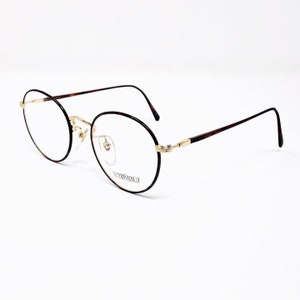 May include: Oval eyeglasses with a thin, dark brown frame and gold-coloured accents. The temples feature a tortoiseshell pattern. The lenses are clear, and the brand name "VERSUS" is visible on one lens.
