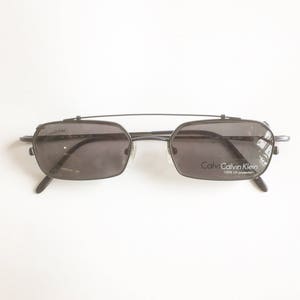 May include: Black rectangular sunglasses with a top bar and the brand name "Calvin Klein" printed on the lens. The sunglasses have dark lenses and a metal frame. The sunglasses are on a white background.