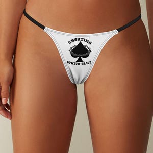 May include: White thong underwear with black trim and the text "CHEATING WHITE SLUT" printed on the front. The design includes a black spade symbol with a crown of thorns.