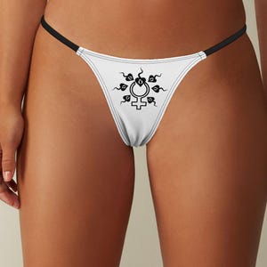 May include: White thong underwear with a black graphic design. The design features a stylized symbol surrounded by decorative elements. The thong has thin black straps and a white fabric front.