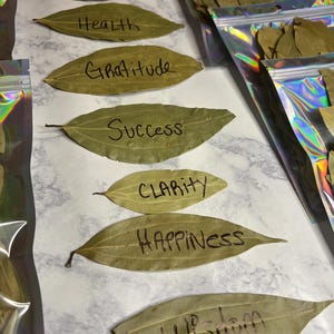 Peut inclure: Feuilles de laurier séchées avec des mots manuscrits : Contentement, Santé, Gratitude, Succès, Clarté, Bonheur et Sagesse. Certaines feuilles sont dans des sacs zippés holographiques. Les feuilles sont de couleur vert clair.