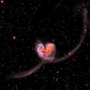 May include: A vibrant image of two galaxies merging, forming a heart-like shape. The galaxies display swirls of red, pink, and blue hues, set against a backdrop of a dark, star-filled cosmos. A curved stream of light extends from the heart shape.