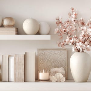 May include: A display of decorative items on white shelves. Includes books, vases, a candle, and a framed picture. A large vase holds pink flowering branches. The colour palette is neutral with soft pink and beige accents.