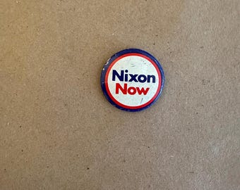 May include: A white button with a blue and red border. The button says "Nixon Now" in red letters.