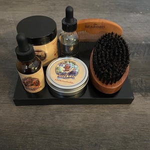 May include: A black tray with a beard oil bottle, beard balm, beard brush, and comb. The beard oil bottle is labeled "King Beard Oil". The beard balm is labeled "The Beard Co. "Villain" Naturally Inspired Beard Balm". The comb is labeled "The Bearded".