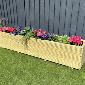 May include: A long wooden planter box with a slatted design. The planter is filled with pink, purple, and green flowers and plants. The planter is sitting on a green lawn in front of a dark grey wooden fence.