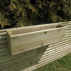 May include: A light-coloured wooden planter box with horizontal slats, attached to a wooden fence. The planter has metal hooks for hanging. The background shows green foliage and a grassy area.