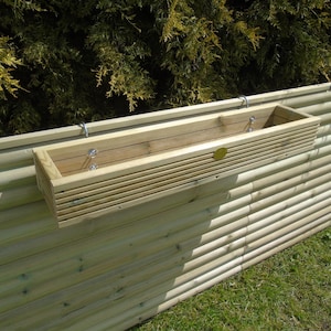 May include: A wooden planter box with a slatted design is mounted on a wooden fence. The planter is made of natural wood and has a light brown colour.