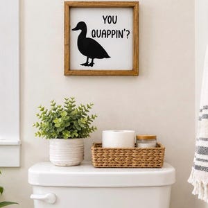 May include: A square wooden framed sign with a white background featuring a black silhouette of a duck and the black text YOU QUAPPIN' in a sans-serif font. The sign is displayed on a wall above a white toilet tank with a plant and basket.