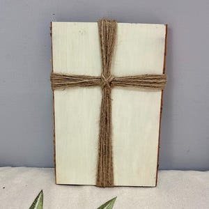 May include: A rectangular wooden plaque with a light-colored surface. A cross made of brown twine is wrapped around the center of the plaque. The edges of the wood have a darker, bark-like appearance. The plaque is a religious item.