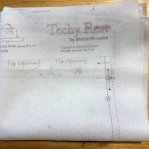 May include: A white sewing pattern for a Techy Rest, a small pillow or cushion, measuring approximately 8 inches by 12 inches. The pattern includes instructions for a top opening and a letter A marking. The text "Techy Rest by Mattie Rhoades" is printed at the top of the pattern.