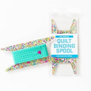 May include: A clear plastic quilt binding spool with a teal spool of binding and a pink heart-shaped pin. The spool is decorated with colorful glitter. The packaging reads "The Original Quilt Binding Spool".