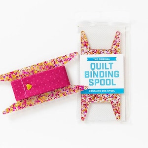 May include: A pink and gold patterned quilt binding spool. The spool is made of plastic and has a pink fabric strip with white polka dots. The packaging says "The Original Quilt Binding Spool" and "Contains One Spool".