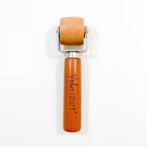 May include: A seam roller with a cylindrical light wood wheel attached to a metal frame and a smooth, cylindrical medium-brown wooden handle. The handle features the text Violet CRAFT engraved vertically in a dark brown script font.