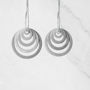 May include: A pair of silver-toned earrings. Each earring features a series of concentric circles, creating a layered, circular design. The earrings hang from thin, curved hooks. The background is a light, neutral color.