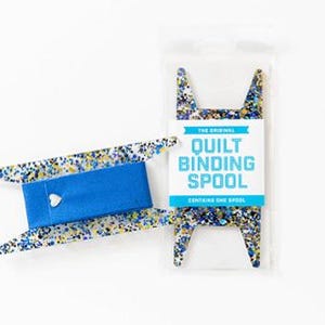 May include: A quilt binding spool with a blue and white speckled design. The spool is packaged in a clear plastic bag with a white label that reads "The Original Quilt Binding Spool". The spool has a blue center with a white heart.