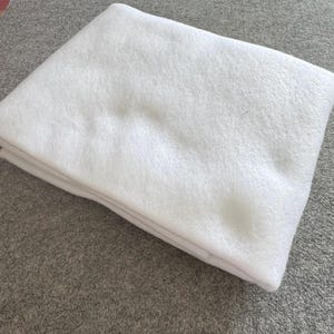 May include: A folded, white, fluffy fabric, possibly a blanket or quilt, is the primary subject. The fabric is soft and appears to be made of a plush material. The background is a neutral gray surface.