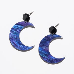 May include: A pair of crescent moon earrings. The earrings have a marbled design in blue and purple, with a black stud. The earrings are set against a white background.