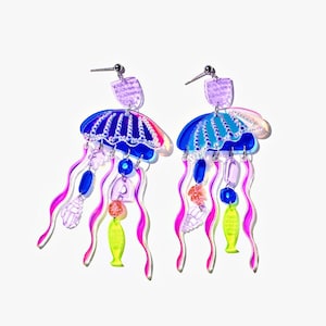 Puede incluir: Un par de pendientes de medusa coloridos. Cada pendiente presenta una parte superior en forma de paraguas azul y rosa con tentáculos iridiscentes colgantes, cuentas azules y rosas y un dije de pez amarillo. Los pendientes tienen postes plateados.