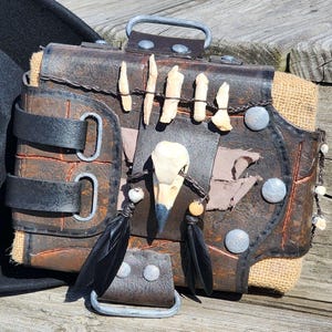 Peut inclure: Un chapeau de cowboy en feutre noir est posé à côté d'un sac en cuir fait à la main. Le sac présente des accents d'os, des clous métalliques et des glands de plumes noires, avec un aspect rustique et vieilli. Le sac a une boucle et des sangles.