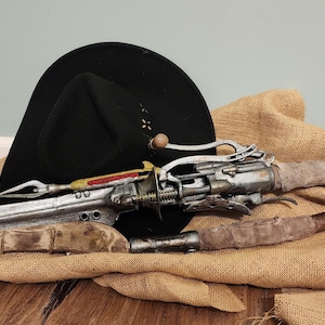May include: A futuristic-looking prop weapon with a silver blade and intricate mechanical details, accented with red and gold. It rests on a burlap cloth next to a black cowboy hat. The weapon has a wrapped handle and a unique design.
