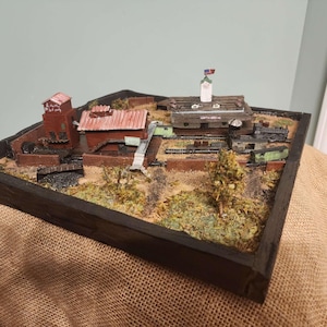 Könnte beinhalten: Miniatur-Diorama einer Stadtszene, eingebettet in einen dunklen Holzrahmen. Die Szene umfasst Gebäude mit roten Dächern, eine Bahngleis und kleine Bäume. Auf einem der Gebäude befindet sich eine Flagge. Das Diorama befindet sich auf einer Jutefläche.