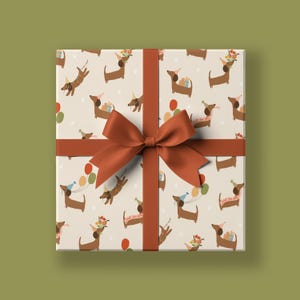 Könnte beinhalten: Eine weiße Geschenkbox mit braunem Band und Schleife. Die Box ist in Geschenkpapier mit einem Muster aus braunen Dackeln eingepackt, die Partyhüte tragen und Ballons halten. Der Text auf dem Geschenkpapier lautet "Happy Paw-ty To You".