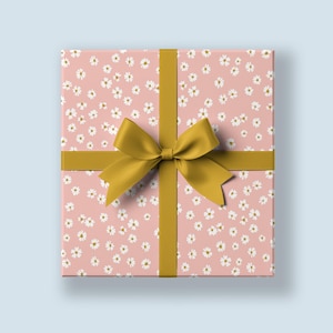 Könnte beinhalten: Eine rosa Geschenkbox mit einem weißen Blumenmuster und einem goldenen Band, das zu einer Schleife gebunden ist.