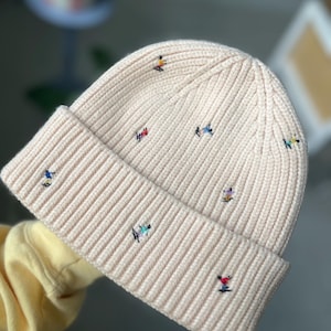 May include: A cream-colored knit beanie with a folded brim. The hat is embroidered with small, colorful skier figures. The beanie is made of a ribbed knit material and is a winter accessory.