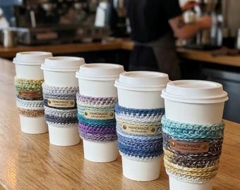 May include: Five white disposable coffee cups arranged in a row on a wooden surface. Each cup features a unique, multi-coloured crocheted sleeve with a small, rectangular brown leather tag embossed with the word HANDMADE attached to the front.