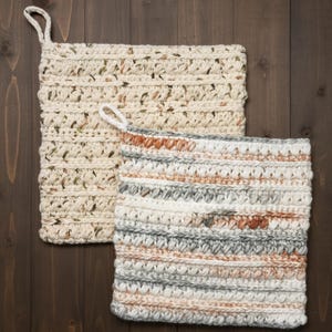 May include: Two handmade crochet potholders. One is cream-coloured with flecks of brown and green. The other is a mix of white, grey, and brown stripes. Both have a loop for hanging. The potholders are square and sit on a dark wood surface.