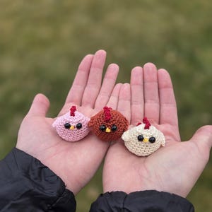 Puede incluir: Tres pollitos de crochet en rosa, marrón y blanco con ojos negros. Los pollitos están sentados en la palma de la mano de una persona.