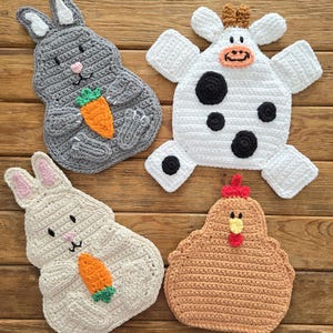 Puede incluir: Cuatro salvamanteles hechos a mano con ganchillo con forma de animales. Incluye un conejito gris con una zanahoria, un conejito blanco, una vaca blanca con manchas negras y una gallina marrón. Sobre una superficie de madera.