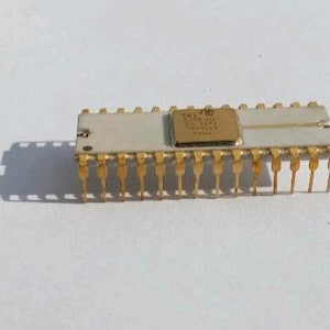Pode incluir: Um circuito integrado retangular com um corpo bege claro e pinos dourados. Um pequeno componente retangular dourado está centrado na parte superior. O componente está sobre um fundo branco.