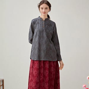 Könnte beinhalten: Eine Frau trägt ein graues Langarm-Oberteil mit floralem Muster und einen roten, bodenlangen Rock mit Blumenmuster. Das Oberteil hat einen Stehkragen und Holzknöpfe. Das Outfit wird durch schwarze Stiefel ergänzt.