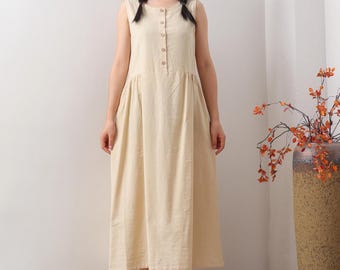 May include: A sleeveless, light beige dress with a button-down front and a gathered skirt. The dress falls to mid-calf length. The woman is wearing beige flats. A tall, textured vase with orange branches is in the background.