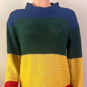Puede incluir: Un suéter de bloques de color con cuello alto. El suéter presenta rayas horizontales en azul, verde, amarillo y rojo. Las mangas son largas y el dobladillo es curvo. El suéter está hecho de punto.