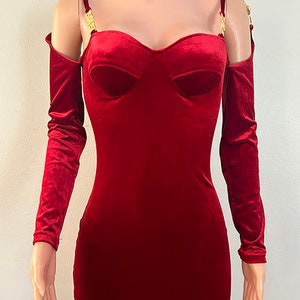 May include: A red velvet dress with long sleeves and a sweetheart neckline. The dress has a fitted bodice and a flowing skirt. The dress features a gold chain detail at the shoulders.