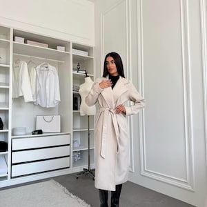 Op de afbeelding: Een lange, lichtbeige jas met bijpassende riem, gedragen met zwarte laarzen. De jas heeft een klassiek ontwerp met een reverskraag. De foto is genomen in een witte kast met een vloerkleed op de vloer.