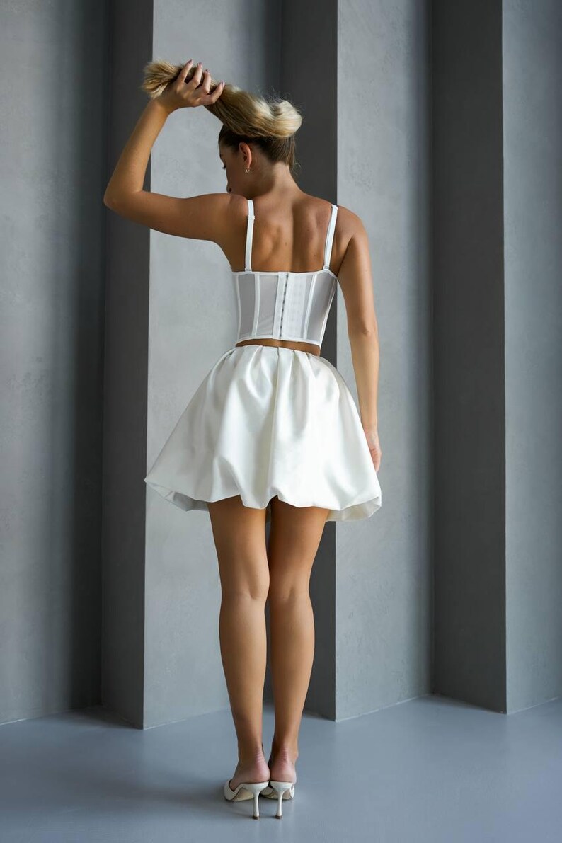 May include: A white corset top with spaghetti straps and a matching mini skirt. The outfit is made of a satin-like material. The model is wearing white heels.