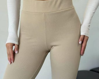 May include: Beige ribbed leggings with a high waist. The leggings are worn with a white long-sleeved crop top. The fabric has a vertical ribbed texture.